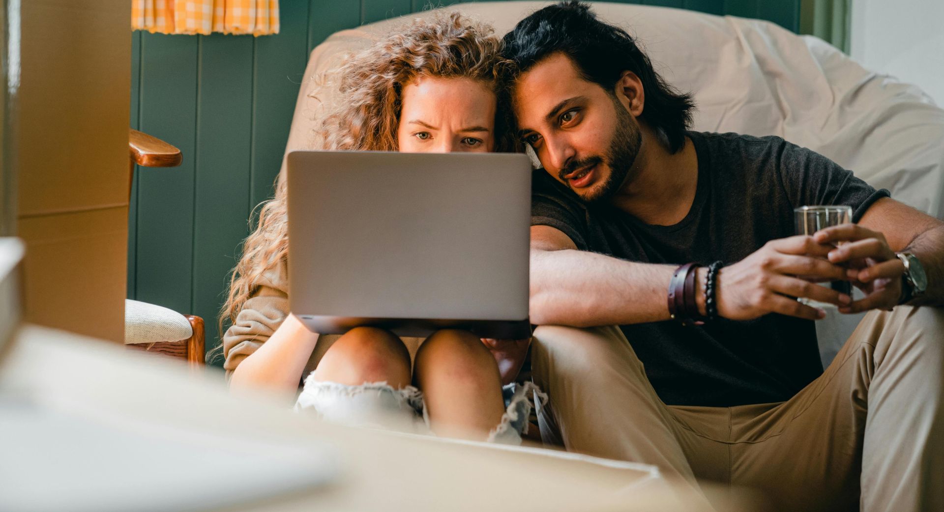 Focused young diverse couple in casual clothes sitting on floor against chair under white cover and browsing netbook